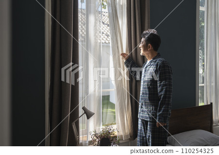 A man in his 50s wearing pajamas opening the curtains in his bedroom A man in his 50s wearing pajamas opening the curtains in his bedroom 110254325
