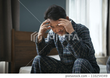 A man in his mid-50s sitting on the bed and holding his head 110254349