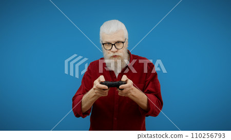 Senior man presses buttons on a joystick against a blue background, embracing his inner video game geek Senior man presses buttons on a joystick against a blue background, embracing his inner video game geek 110256793