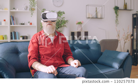 A senior man wears virtual reality glasses while seated on a sofa, exploring the world through a VR headset A senior man wears virtual reality glasses while seated on a sofa, exploring the world through a VR headset 110257078