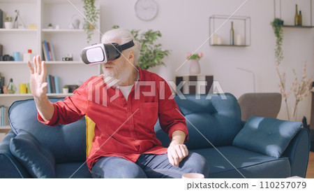 A mature man experiences virtual reality, seated on a sofa with a VR headset, as he reaches out into virtual space A mature man experiences virtual reality, seated on a sofa with a VR headset, as he reaches out into virtual space 110257079