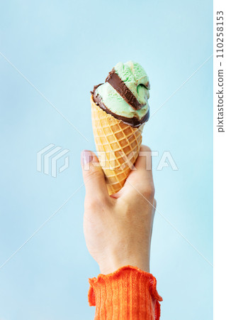 A hand holding an ice cream cone with green and brown scoops against a light blue background. 110258153