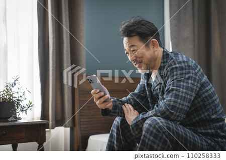 微笑的中年男子坐在床上,看著智慧型手機 微笑的中年男子坐在床上,看著智慧型手機 110258333
