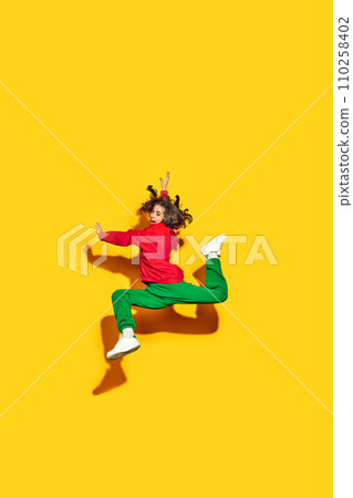 Fashion takes flight. Young happy woman, student dressed in trendy, modern sportwear and hat jumping in motion against yellow background. 110258402