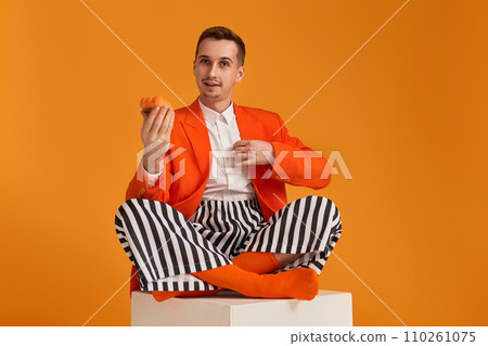 extravagant funny young man in orange jacket and striped pants holding cupcake on orange background. extravagant funny young man in orange jacket and striped pants holding cupcake on orange background. 110261075