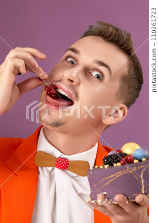 handsome birthday man in orange jacket eating cake on purple background 110261723