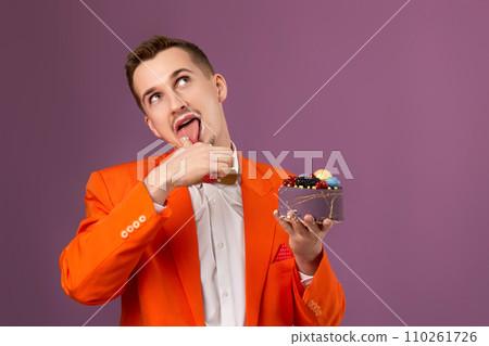 happy man in orange jacket eating cream cake on purple background 110261726