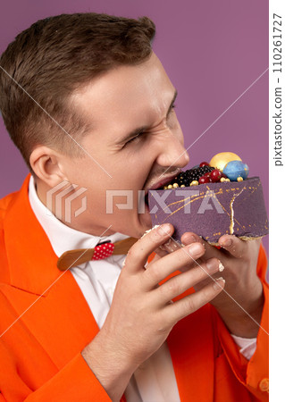 handsome birthday man in orange jacket bites cake on purple background 110261727