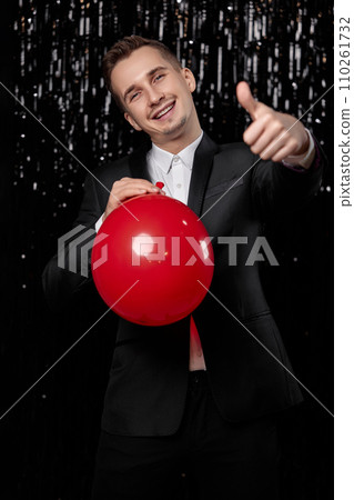caucasian smiling man in black jacket with red air balloon showing ok gesture on dark glitter background. 110261732
