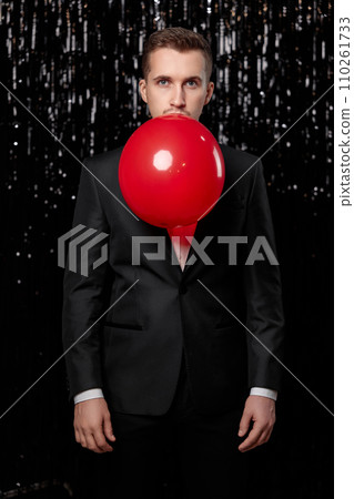 caucasian man in black jacket blowing red air balloon on glitter background. new year party 110261733