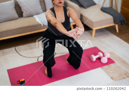 Overweight woman enjoying a fitness workout at home. Fat, plump woman and squatting on an exercise mat in the living room Overweight woman enjoying a fitness workout at home. Fat, plump woman and squatting on an exercise mat in the living room 110262042