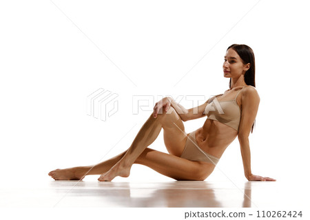 Beautiful young woman with relief slim fit body sitting on floor, posing in beige cotton underwear against white studio background 110262424