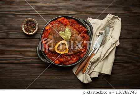 Traditional Italian dish Ossobuco all Milanese made with cut veal shank meat with vegetable tomato sauce served in black casserole pan top view on rustic brown wooden background Traditional Italian dish Ossobuco all Milanese made with cut veal shank meat with vegetable tomato sauce served in black casserole pan top view on rustic brown wooden background 110262746