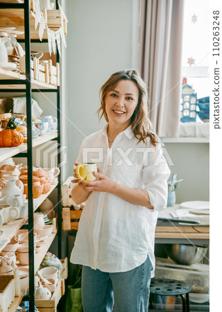 A beautiful middle-aged woman stands at shelf with various pottery items. A young woman presents clay products in her shop-workshop, small business. 110263248