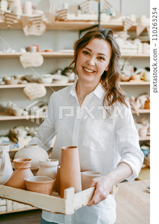 A female potter holds a box with raw clay products. An Artisan woman happily demonstrates her handicrafts in a pottery workshop A female potter holds a box with raw clay products. An Artisan woman happily demonstrates her handicrafts in a pottery workshop 110263254