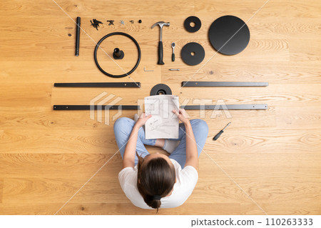 A woman with instructions is sitting among furniture parts laid out on the floor 110263333