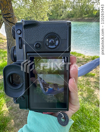 On the camera's display, a man is resting on a hammock in the open air near a river. On the camera's display, a man is resting on a hammock in the open air near a river. 110263443