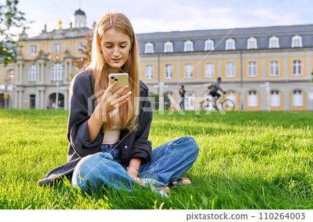 Young smiling female using smartphone sitting on grass, lawn in sunset light 110264003