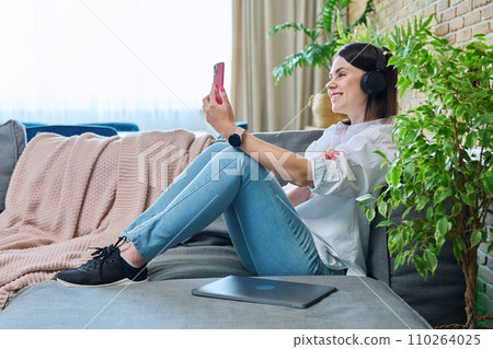 Young woman having video call sitting on sofa at home using smartphone 110264025