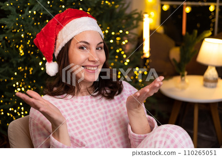 A young attractive woman in cozy pajamas is sitting at home on a chair in a red Christmas hat, smiling and spreading her arms to the sides A young attractive woman in cozy pajamas is sitting at home on a chair in a red Christmas hat, smiling and spreading her arms to the sides 110265051