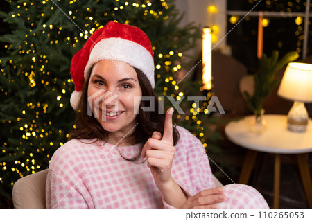 An attractive young woman in cozy pajamas is sitting at home on a chair in a red Christmas hat, smiling and holding her index finger up. An attractive young woman in cozy pajamas is sitting at home on a chair in a red Christmas hat, smiling and holding her index finger up. 110265053