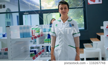 Portrait of friendly pharmacist in apothecary placing cardiovascular pills box on shelves. Smiling healthcare professional in drugstore arranging medicinal merchandise, ready to assist clients 110267748