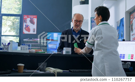Older shopper arriving at drugstore checkout counter after finding needed medical products, purchasing fever stopping pills with credit card, being helped by chemist assistant with shopping bag Older shopper arriving at drugstore checkout counter after finding needed medical products, purchasing fever stopping pills with credit card, being helped by chemist assistant with shopping bag 110267757