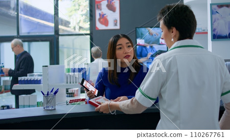 Asian customer at pharmacy counter, showing druggist prescribed heart medicine and inquiring about required dosage. Healthcare professional assisting client with needed medicinal product Asian customer at pharmacy counter, showing druggist prescribed heart medicine and inquiring about required dosage. Healthcare professional assisting client with needed medicinal product 110267761