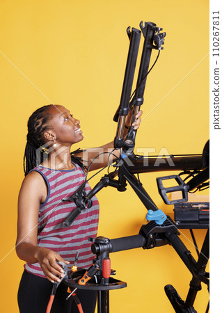 Healthy active black woman repairs bike damage with professional repair-stand and tools against isolated yellow background. Athletic female examining and fixing bicycle with specialized equipment. 110267811