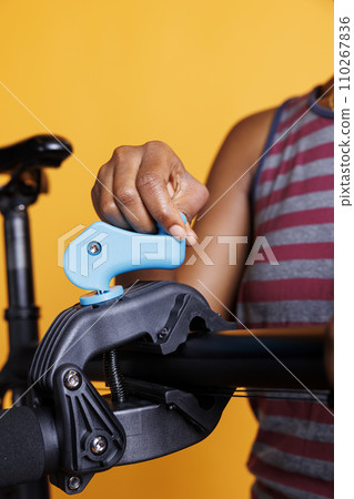 African american woman tightening clamp to secure bicycle on repair stand for adjustments. Close-up shot of female cyclist placing a damaged bike on a workstand for maintenance. African american woman tightening clamp to secure bicycle on repair stand for adjustments. Close-up shot of female cyclist placing a damaged bike on a workstand for maintenance. 110267836
