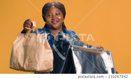 While delivering takeout meals on bicycle, youthful black woman giving thumbs up to ensure client satisfaction. In local neighborhood african american delivery person provides eco-friendly dine-in 110267842