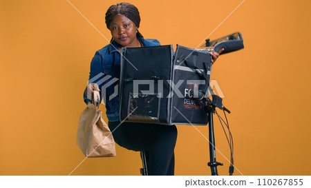 African american woman grabbing takeaway from food delivery bag for person in local neighborhood. Smiling courier providing fast and reliable customer service by giving fresh takeout meal to client. 110267855