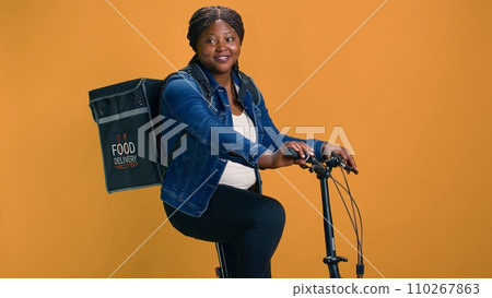Young and active courier using modern bicycle to deliver package with precision to the city office. African american lady with backpack working as professional delivery person. Young and active courier using modern bicycle to deliver package with precision to the city office. African american lady with backpack working as professional delivery person. 110267863