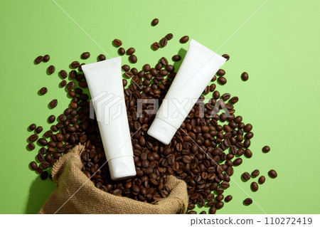 On a green background, coffee beans falling from a burlap bag and two empty cosmetic tubes are decorated. Top view, mockup scene for advertising. Natural ingredient for skin care and body care On a green background, coffee beans falling from a burlap bag and two empty cosmetic tubes are decorated. Top view, mockup scene for advertising. Natural ingredient for skin care and body care 110272419