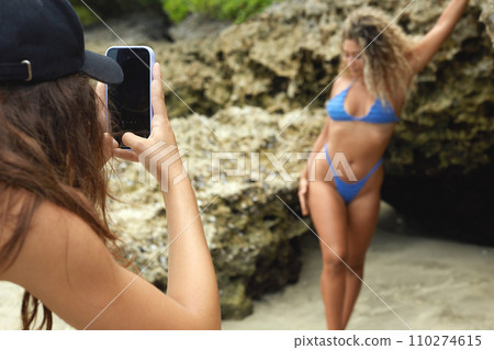 photo shoot on the phone. model in bikini posing for photographer. two girls taking pictures on the beach 110274615