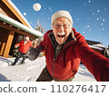 Joyful elderly man in a red jacket takes a selfie while playfully throwing a snowball. Another man in a Santa hat is in the background in a snowy setting 110276417