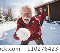 Joyful elderly man in a red jacket takes a selfie while playfully throwing a snowball. Another man in a Santa hat is in the background in a snowy setting 110276423