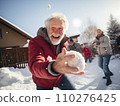 Joyful elderly man in a red jacket takes a selfie while playfully throwing a snowball. Another man in a Santa hat is in the background in a snowy setting 110276425