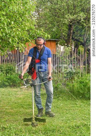 A male gardener mows the green grass in the backyard with a gasoline mower. care of a garden plot A male gardener mows the green grass in the backyard with a gasoline mower. care of a garden plot 110276770