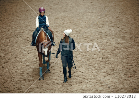 Aerial, top view. Female teacher, instructor teaching little girl, kid horseback riding. Careful ride, outdoor lesson Aerial, top view. Female teacher, instructor teaching little girl, kid horseback riding. Careful ride, outdoor lesson 110278018