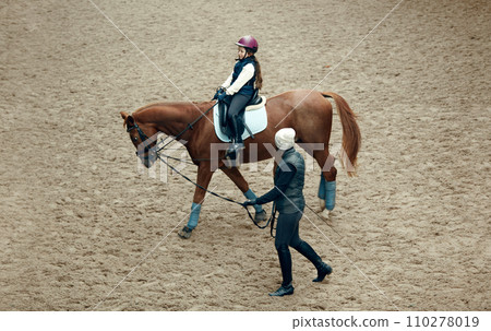 Aerial, top view. Female teacher, instructor teaching little girl, kid horseback riding. Careful ride, outdoor lesson Aerial, top view. Female teacher, instructor teaching little girl, kid horseback riding. Careful ride, outdoor lesson 110278019