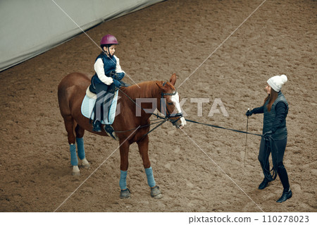 Aerial, top view. Female teacher, instructor teaching little girl, kid horseback riding. Outdoor lesson Aerial, top view. Female teacher, instructor teaching little girl, kid horseback riding. Outdoor lesson 110278023