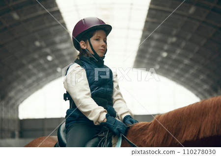 Portrait of little girl, child in helmet sitting on brown horse, training, practicing horseback riding. 110278030