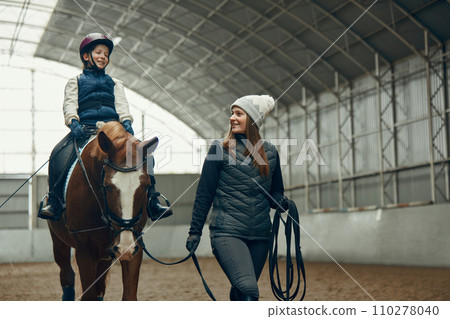 Young woman, teacher, instructor teaching little girl horseback riding. Child sitting in paddle, practicing. Education 110278040