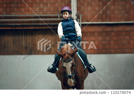 Little girl, child in uniform and helmet sitting on horse, raining, learning horseback riding activity on special arena, pavilion Little girl, child in uniform and helmet sitting on horse, raining, learning horseback riding activity on special arena, pavilion 110278074