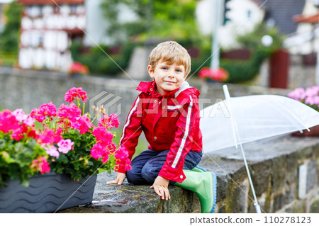 Little blond kid boy walking with big umbrella outdoors on rainy day. Portrait of cute preschool child having fun wear colorful waterproof clothes. Outdoor leisure walk on bad weather day with kids 110278123