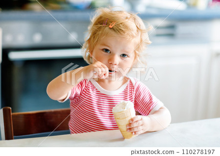 Adorable baby girl eating from spoon sweet ice cream in waffle cone. food, child, feeding and development concept. Cute toddler, daughter with spoon sitting in highchair and learning eat by itself 110278197