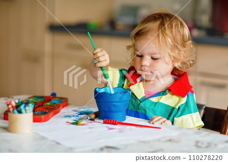 Cute adorable baby girl learning painting with water colors. Little toddler child drawing at home, using colorful brushes. Healthy happy daughter experimenting with colors, water at home or nursery. 110278220