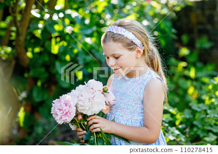 Cute adorable little preschool girl with huge bouquet of blossoming pink peony flowers. Portrait of smiling preschool child in domestic garden on warm spring or summer day. Summertime. 110278415
