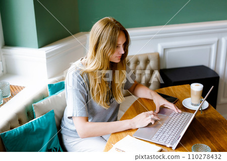 Portrait of young woman using technology of smartphone and looking at laptop. Young woman freelancer using laptop computer working in restaurant or indoor cafe with coffee. Home office concept. 110278432